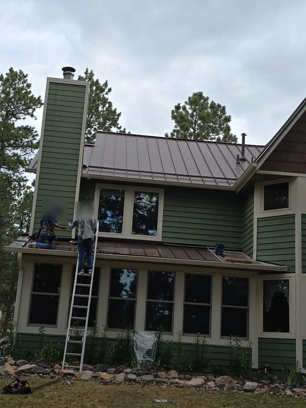 Two roofers on ladders at the eave of a two-story home with a brown standing seam metal roof