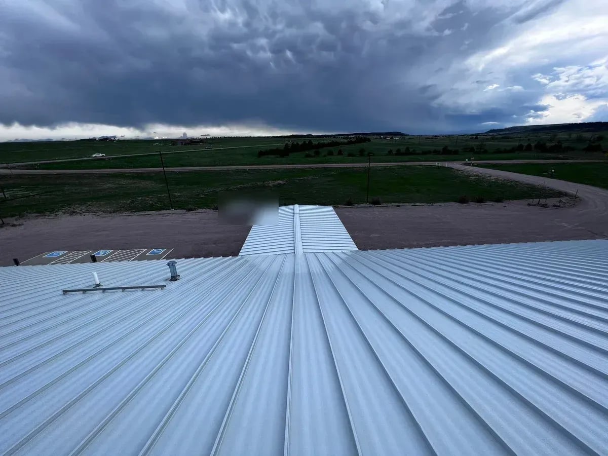 Long silver standing seam metal roof on a large building with open fields under a stormy sky